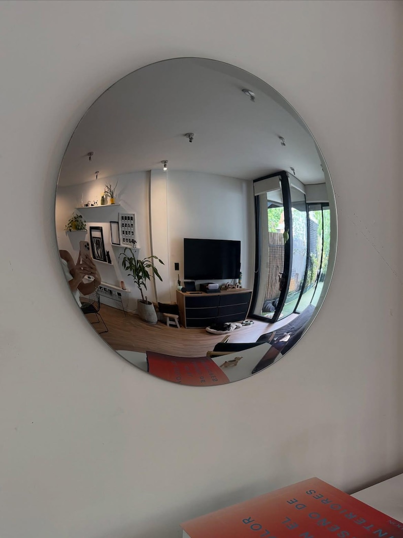 May include: A large, circular, frameless wall mirror with a reflective surface that creates a wide-angle, convex-style distortion of the room it faces, showing a living area with a television, furniture, and indoor plants.