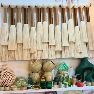 May include: A wall hanging featuring a horizontal wooden rod with multiple vertical strands of cream-colored yarn, each wrapped in alternating brown and tan thread. Below, a shelf displays various ceramic vases, green glass cups, and woven baskets.
