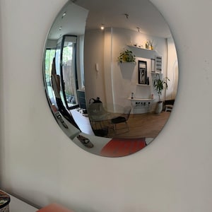 May include: A large, circular, frameless convex mirror mounted on a wall. The reflective surface creates a wide-angle, distorted view of an interior room, including furniture, plants, and a person, while reflecting a book with the text EL COLOR EN EL DISE&Ntilde;O DE INTERIORES.