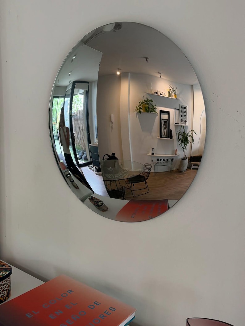 May include: A large, circular, frameless convex mirror mounted on a wall. The reflective surface creates a wide-angle, distorted view of an interior room, including furniture, plants, and a person, while reflecting a book with the text EL COLOR EN EL DISE&Ntilde;O DE INTERIORES.