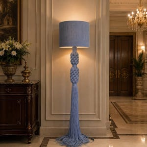 Puede incluir: Una lámpara de pie con una pantalla cilíndrica de tela azul claro y una columna central compuesta por dos secciones de cuerda azul anudadas de forma intrincada. La base de la lámpara termina en largos flecos azules que se extienden por el suelo.