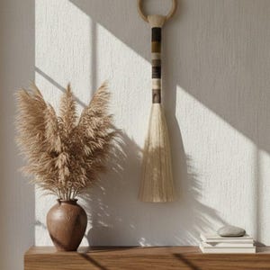 May include: A decorative wall hanging with a circular top and a striped handle in shades of brown, beige, and olive green. The hanging is made of natural fibers and hangs next to a vase with dried pampas grass. A wooden shelf holds books and a stone.