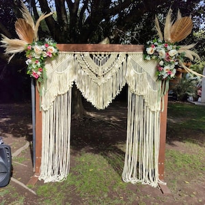 Op de afbeelding: Een houten boog versierd met een macramé wandkleed en bloemstukken. De boog is gemaakt van natuurlijk hout en heeft een rustieke uitstraling. Het macramé wandkleed is wit en heeft een gefranst ontwerp. De bloemstukken zijn gemaakt van witte en roze bloemen en groen.