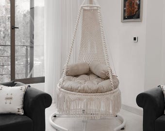 May include: A hanging macrame chair with a cream-colored cushion and two matching pillows. The chair is suspended from the ceiling and has a white metal frame. The chair is in a bright, modern living room setting.