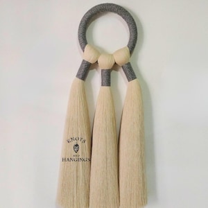 May include: A handcrafted wall hanging featuring a gray circular top with three cream-colored tassels. The tassels are connected to the top with gray and cream-colored accents. The text "KNOTS AND HANGINGS" is printed on one of the tassels.