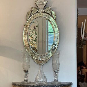 May include: An oval-shaped, ornate, silver-toned mirror with a decorative leaf design. The mirror is hanging above a marble console table with two crystal candle holders and a crystal decanter.