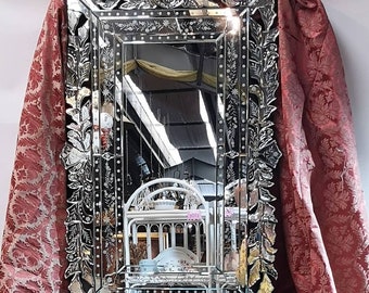 May include: Rectangular mirror with an ornate, etched frame. The frame features floral and leaf designs, with a decorative crown at the top. The mirror reflects a room with white furniture and a red patterned curtain.