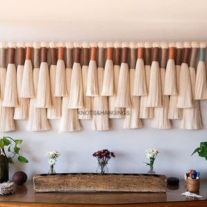 Könnte beinhalten: Ein Wandbehang aus weißem Garn mit braunen, orangen und blauen Akzenten. Der Behang besteht aus mehreren Garnsträngen, die zu einem Fransen-Stil zusammengebunden sind. Der Behang wird an einer Wand über einem Holzregal mit Dekorationsartikeln gezeigt.