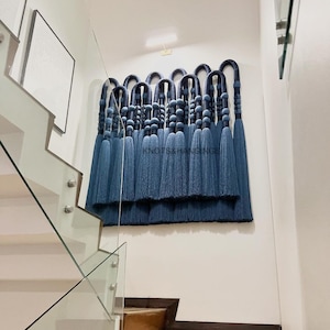 May include: A wall hanging made of blue and navy blue yarn with a fringe. The hanging is made of multiple strands of yarn that are knotted together to create a wave-like pattern. The hanging is titled "Knots & Hangs".