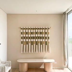 May include: A decorative wall hanging featuring a series of white, spherical beads and tassels. The beads are interspersed with green and gold accents, and the tassels hang below. The piece is mounted on a wooden rod, creating a unique wall art piece.