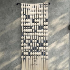 May include: A large, off-white wall hanging with a repeating pattern of white, oval-shaped beads and vertical sections of blue and white woven material. The bottom of the hanging has long, white tassels. The text "Knots and Hangings" is visible.