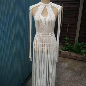 May include: A cream-colored macrame dress displayed on a mannequin. The dress features a halter neckline with a keyhole detail, a fitted waist, and long fringe. The dress is made of woven cords and has long fringe that hangs down to the floor.