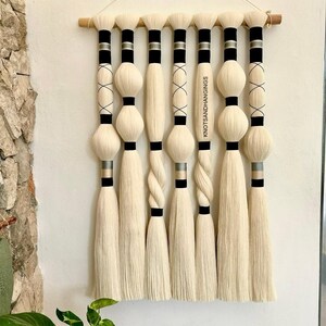 May include: A wall hanging made of white yarn with black and gold accents. The yarn is woven into a series of tassels and knots. The wall hanging is hung on a wooden dowel. The text "KnotsAndHangings" is visible on the right side of the wall hanging.