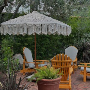 Puede incluir: Una sombrilla de patio de macramé color crema con un poste de madera y bordes con flecos. La sombrilla está instalada en un jardín con sillas de madera, un patio de ladrillos y una maceta de terracota.