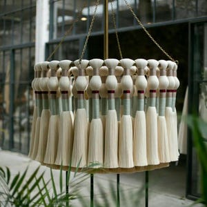 May include: A decorative chandelier featuring a circular design with multiple off-white tassels. The tassels are arranged in a layered pattern, suspended from a brass-colored frame and chain. The chandelier is set against a backdrop of windows and greenery.