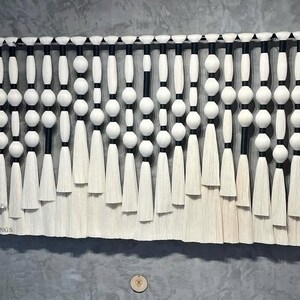 May include: A decorative wall hanging featuring a black rod with white beads and tassels. The design includes white spheres and elongated shapes, with black accents. The bottom edge has a fringe-like texture. The text "KNOT" and "HANGINGS" is visible.