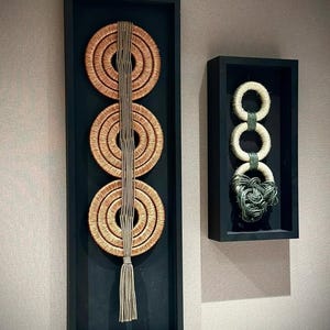 May include: Two framed wall art pieces. One features three woven, circular designs in a vertical black frame, connected by a central cord and tassel. The other has three white circular designs with a green knotted accent, in a black frame.