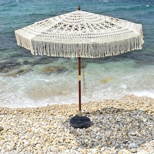 Peut inclure: Un parasol de plage en crochet crème avec une bordure à franges se dresse sur une plage de galets. Le parasol a un mât en bois et une base circulaire foncée. L'océan est visible en arrière-plan.