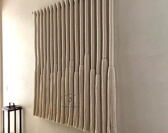 May include: A large, beige macrame wall hanging with multiple vertical strands, each ending in a tassel. The artwork is mounted on a light-coloured wall. A dark lantern with a candle sits to the left.