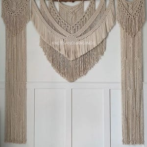 May include: A set of five macrame wall hangings in a cream colour. The hangings are made of cotton cord and feature a variety of knots and patterns. The hangings are arranged in a staggered pattern on a white wall. The text "Knots & Hangings" is visible in the centre of the image.