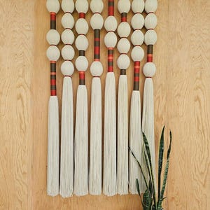 May include: A large, cream-colored macrame wall hanging with a wooden top. The design features rows of cream-colored spheres and long tassels, accented with red and brown stripes. Two potted plants are in the lower right corner.