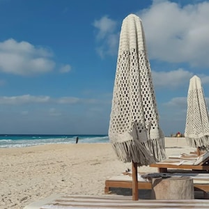 Può includere: Scena sulla spiaggia con due ombrelloni chiusi all'uncinetto color crema con frange. Gli ombrelloni sono su pali di legno, con sedie a sdraio davanti. La sabbia è bianca, l'oceano è blu e il cielo è azzurro con nuvole.