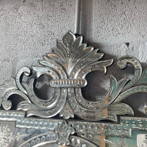 May include: A large, ornate, silver-toned, beveled mirror with a decorative, floral design. The mirror has a large, central oval shape with a smaller, rectangular shape above it. The mirror is framed with a decorative, floral design.