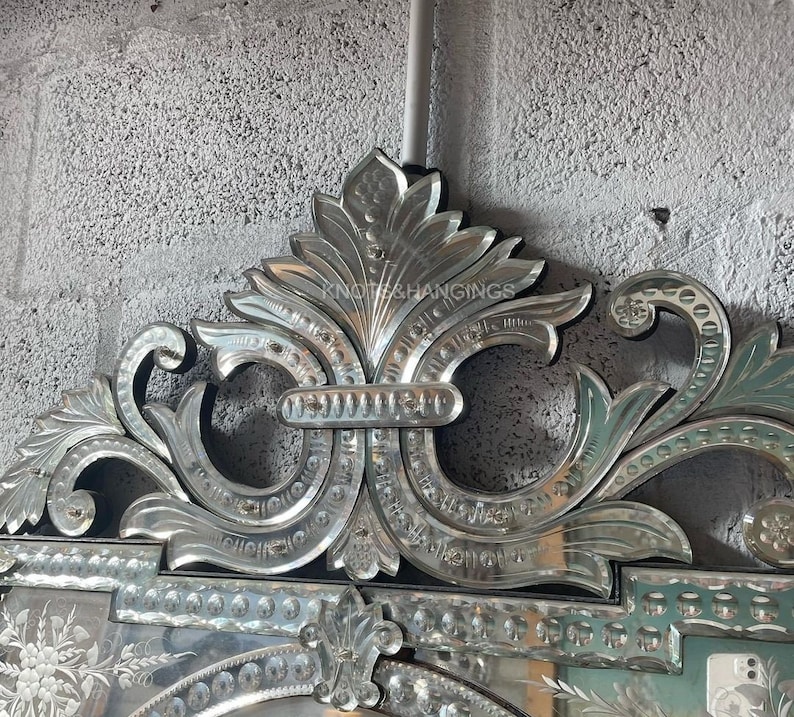 May include: A large, ornate, silver-toned, beveled mirror with a decorative, floral design. The mirror has a large, central oval shape with a smaller, rectangular shape above it. The mirror is framed with a decorative, floral design.