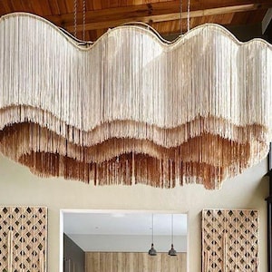 Könnte beinhalten: Ein großer, mehrschichtiger Kronleuchter mit wellenförmigem Design. Die Leuchte besteht aus cremefarbenen Fransen mit einem hellbraunen Farbverlauf am unteren Rand. Sie hängt in einem modernen Wohnraum von der Decke und verleiht ihm einen Hauch von Bohème-Stil.