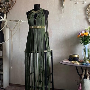 Op de afbeelding: Olijfgroene macramé jurk op een zwarte paspop. De jurk heeft een halternek, een goudkleurige band in de taille en lange franjes. De jurk wordt gepresenteerd op een zwarte metalen standaard.