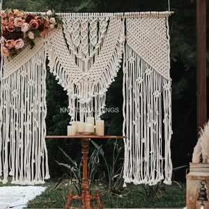 May include: A white macrame wedding backdrop with a geometric pattern. The backdrop is made of cotton rope and is hung from a wooden beam. There are candles on a table in front of the backdrop.