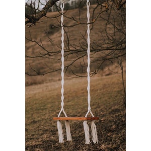 May include: A white rope swing with a wooden seat hangs from a tree branch. The swing has long tassels at the bottom.