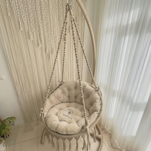 Peut inclure: Un fauteuil suspendu en macramé beige avec un coussin touffeté et des franges. La chaise est suspendue à un cadre métallique incurvé. Le coussin porte le texte "Knots & Hangings". Thème de décoration bohème.