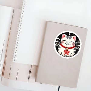 May include: A white and gray lucky cat with a red collar and a gold bell, sitting in a circle with a black background. The cat has a smiling face and a red flower on its chest.