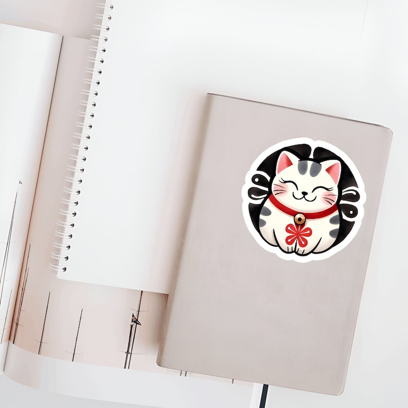 May include: A white and gray lucky cat with a red collar and a gold bell, sitting in a circle with a black background. The cat has a smiling face and a red flower on its chest.