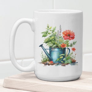 Puede incluir: Taza de cer&aacute;mica blanca con una regadera azul llena de flores y vegetaci&oacute;n. Las flores son rojas, rosas y moradas. La regadera tiene un pico y un asa. La taza est&aacute; sobre una superficie de madera.