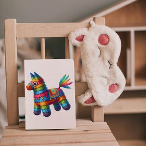 May include: A white notebook with a colorful pinata donkey illustration on the cover. The notebook is sitting on a wooden chair with a white fuzzy eye mask hanging on the back of the chair.