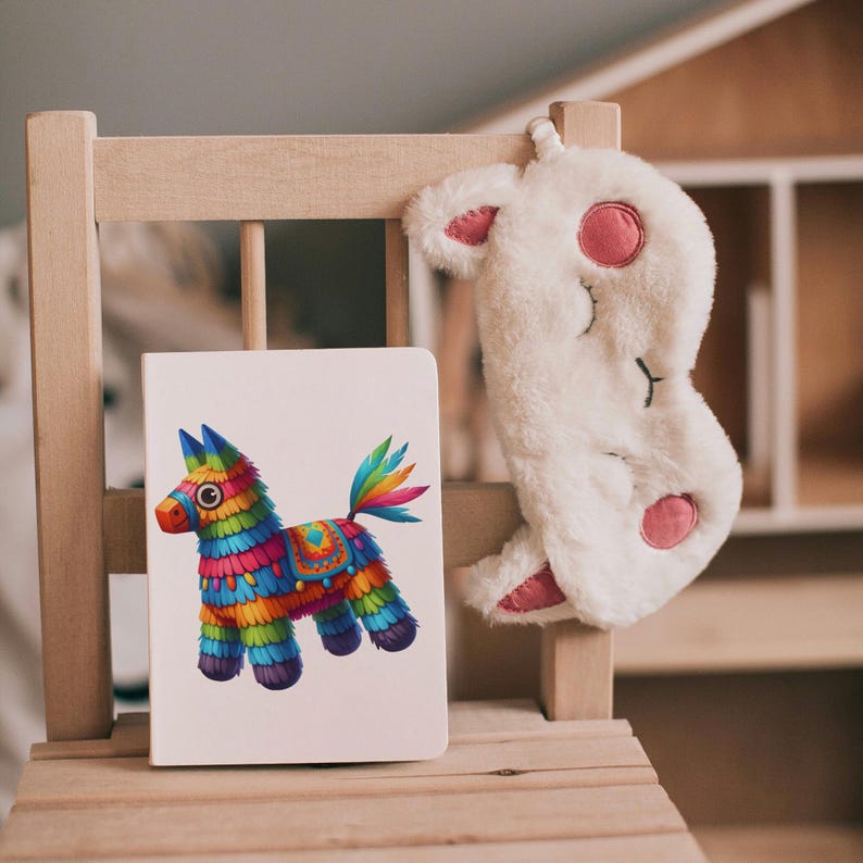 May include: A white notebook with a colorful pinata donkey illustration on the cover. The notebook is sitting on a wooden chair with a white fuzzy eye mask hanging on the back of the chair.