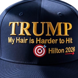 May include: Navy blue baseball cap with gold embroidered text reading "TRUMP" above the phrase "My Hair is Harder to Hit". A target graphic and "Hilton 2026" are also embroidered.