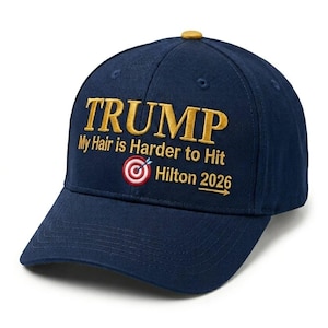 May include: Navy blue baseball cap with the word "TRUMP" embroidered in gold. The cap features the text "My Hair is Harder to Hit" and "Hilton 2026" with a target graphic. The cap has a curved brim and a button on top.