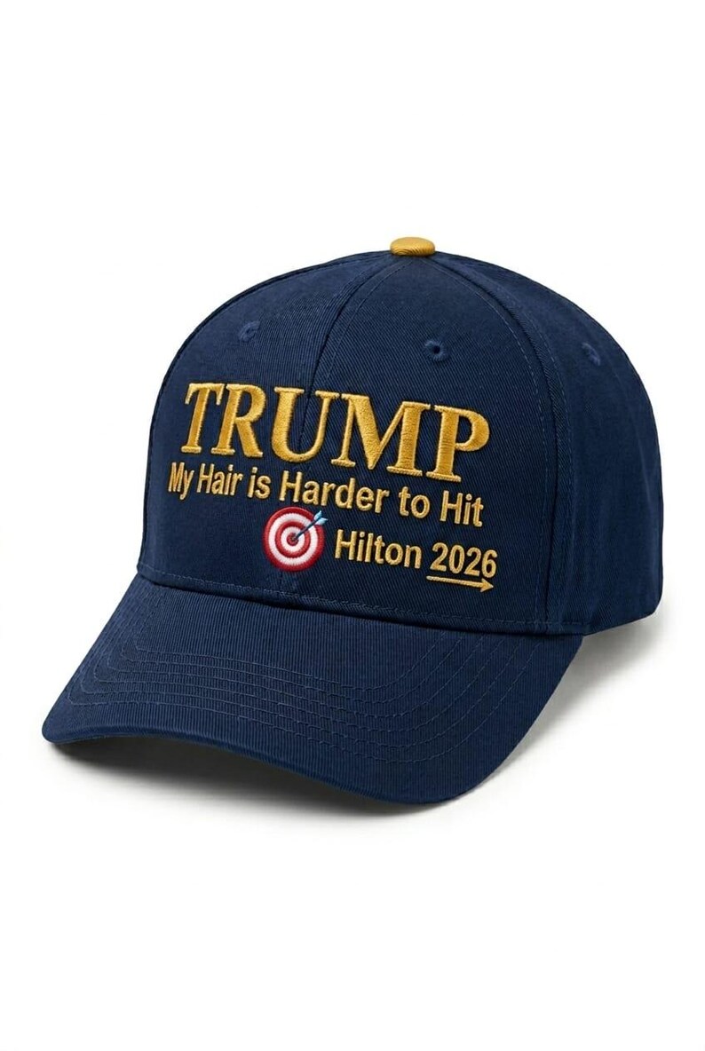 May include: Navy blue baseball cap with the word "TRUMP" embroidered in gold. The cap features the text "My Hair is Harder to Hit" and "Hilton 2026" with a target graphic. The cap has a curved brim and a button on top.