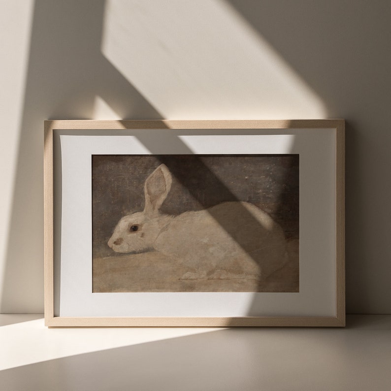 Puede incluir: Una pintura enmarcada de un conejo blanco con ojos marrones, acostado sobre un fondo marr&oacute;n claro. El marco es de madera clara.