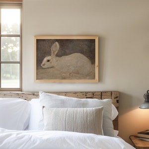 Puede incluir: Una pintura enmarcada de un conejo blanco sentado sobre un fondo marr&oacute;n. La pintura est&aacute; colgada en una pared sobre una cama con ropa de cama blanca y una almohada beige.