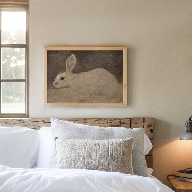 Puede incluir: Una pintura enmarcada de un conejo blanco sentado sobre un fondo marr&oacute;n. La pintura est&aacute; colgada en una pared sobre una cama con ropa de cama blanca y una almohada beige.