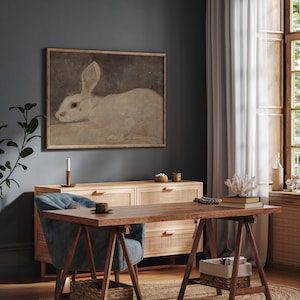 Puede incluir: Una pintura enmarcada de un conejo blanco con orejas largas cuelga de una pared azul oscuro. La pintura est&aacute; sobre una c&oacute;moda de madera con tres cajones. Una mesa de madera con una silla azul est&aacute; delante de la c&oacute;moda.