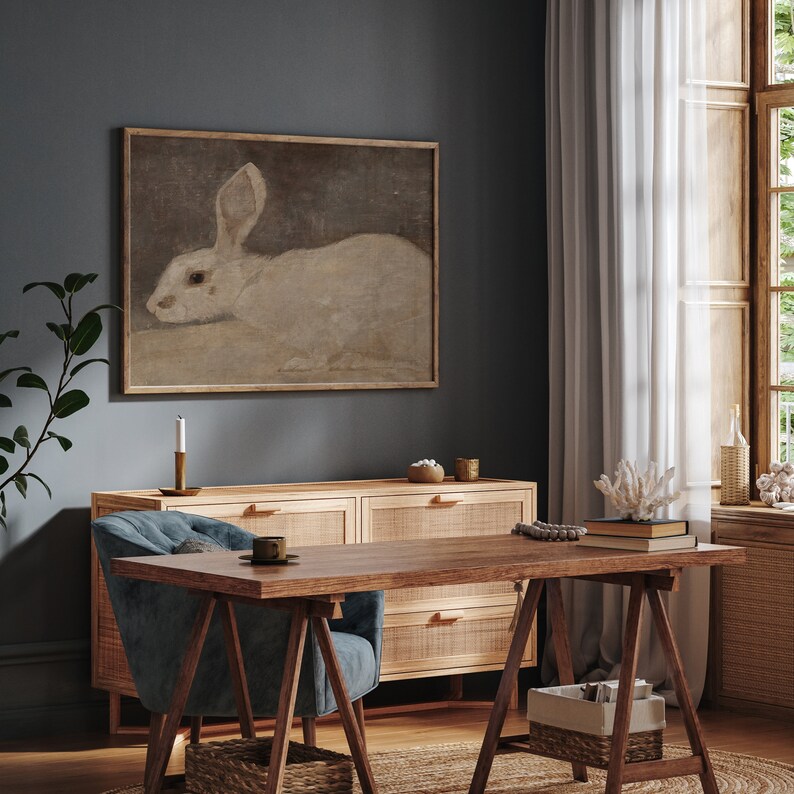 Puede incluir: Una pintura enmarcada de un conejo blanco con orejas largas cuelga de una pared azul oscuro. La pintura est&aacute; sobre una c&oacute;moda de madera con tres cajones. Una mesa de madera con una silla azul est&aacute; delante de la c&oacute;moda.
