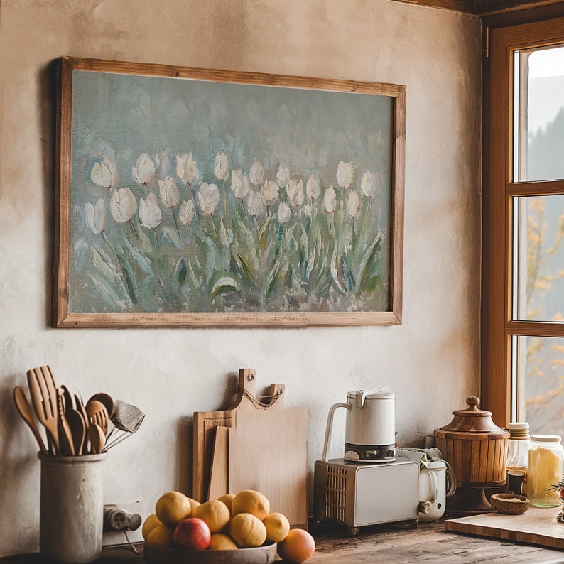 May include: A framed painting of white tulips against a soft blue backdrop hangs above a kitchen counter. The painting is in a rustic wooden frame. Kitchen utensils, cutting boards, and a bowl of fruit are on the counter.