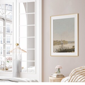 May include: A framed landscape painting with a gold frame. The painting depicts a lake with a sailboat in the distance and trees on the shore. The painting is hanging on a light pink wall.