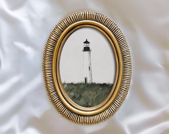May include: An oval, gold-colored framed print featuring a lighthouse. The lighthouse is white with a black top, set against a gray sky and green grass. The frame has a textured, ridged design. The text "ATLAS VINTAGE Curated Vintage Art" is at the bottom.
