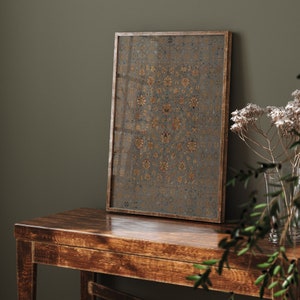 May include: A framed print of a vintage rug with a geometric pattern in shades of gold, blue, and red. The print is hanging on a dark green wall above a wooden table.